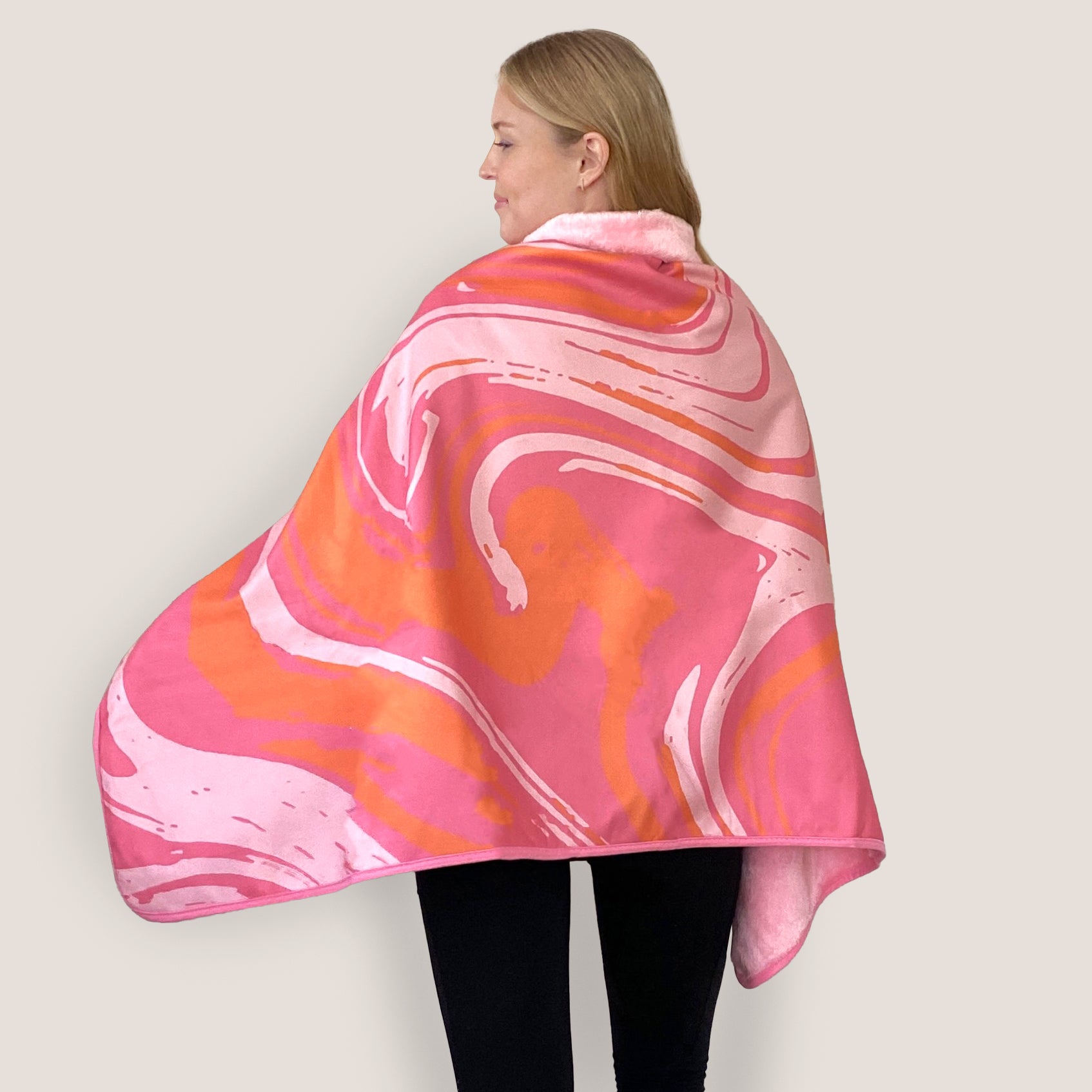 A woman wearing a pink swirls print fleece cape blanket, draping comfortably over her shoulders. The stylish and cozy wrap features a marbled pink and orange pattern, ideal for warmth and fashion.